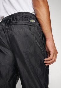 Čierne ľahké joggingové nohavice s diagonálnym diamantovým vzorom, elastickým pásom, bočnými vreckami a malým logom Lacoste na zadnej strane.