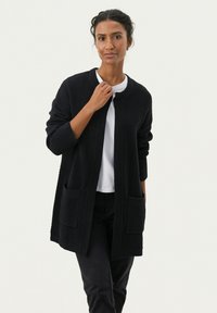 Stickad cardigan i mörk marinblått med ett strukturerat mönster, rund halsringning, öppet fram, två framfickor och långa ärmar. Bärs över en vit topp.