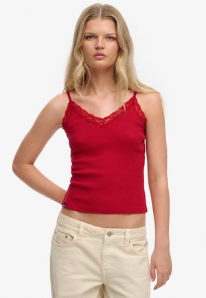 Mujer rubia con top sin mangas rojo con encaje y pantalones beige claros, de pie con una mano descansando detrás de la espalda.