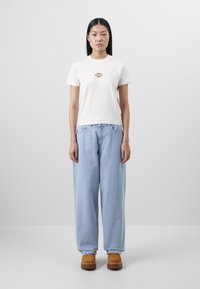 Hvit t-skjorte med kort erme og liten flerfarget logo på brystet, sammen med lys blå baggy jeans og brune klumper.