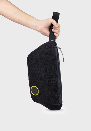 Bolsa de tote negra con textura, exterior suave, detalles de cuero, cierre con cremallera y un logotipo circular amarillo brillante en la parte frontal.