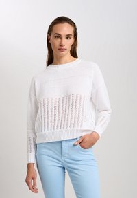 Weißer Strickpullover mit einem verkürzten Design, das eine Mischung aus offenen und festen Mustern aufweist. Rippbündchen und -saum, kombiniert mit hellblauen Hosen.