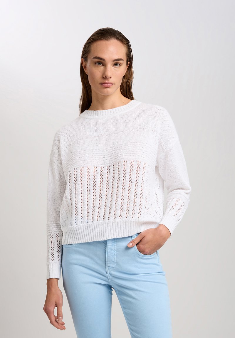 Weißer Strickpullover mit einem verkürzten Design, das eine Mischung aus offenen und festen Mustern aufweist. Rippbündchen und -saum, kombiniert mit hellblauen Hosen.