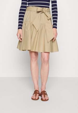 Woman wearing beige A-line skirt with wide tied belt, navy and white striped long-sleeve top, and brown open-toe sandals standing on neutral background.