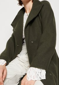 Olive green wool-blend coat with a notched collar and button front, featuring lace-trimmed white sleeves and smooth texture.