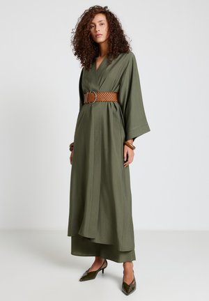 Frau mit lockigem Haar, die ein olivgrünes, langes Kleid mit weiten Ärmeln trägt, dazu einen braunen geflochtenen Gürtel, passende Schuhe und Armbänder, stehend vor weißem Hintergrund.