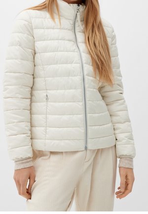 Kobieta ubrana w jasnobeżową pikowaną kurtkę na zamek z bocznymi kieszeniami oraz kremowe, prążkowane spodnie na jednolitym tle.