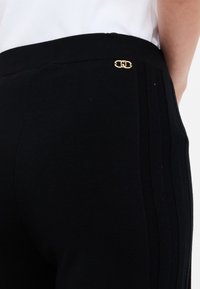 Pantaloni neri in maglia con trama a coste, caratterizzati da un logo con accento dorato sulla vita e dettagli a strisce verticali sui lati.