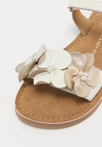 Bela usnjena sandal s cvetličnimi okrasitvami v krem, zlati in srebrni barvi. Udoben rjav sued insole s podrobnostmi v rožnati barvi.