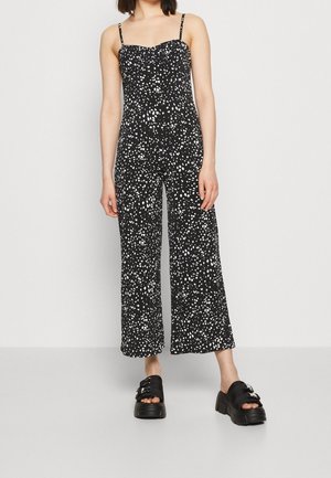 Femme portant une combinaison sans manches noire avec des taches blanches irrégulières et des sandales à plateforme noires avec des boucles en argent, se tenant contre un mur uni.