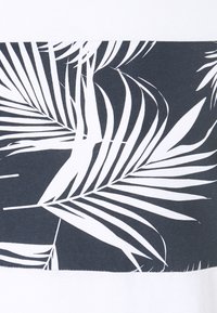 Balts un tumši zils krekls ar palmu lapu rakstu. Audums ir gluds, ar kontrastējošām daļām, veidojot mūsdienīgu dizainu.