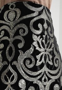 Jupe en velours noir ornée de broderies en sequins argentés, avec des motifs floraux et en spirale, apportant texture et détails au design.