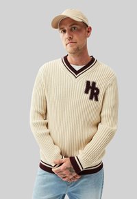 Krämfärgad ribbstickad tröja med maroon-accenter och en "HR"-logotyp. Har en V-hals och åtsittande ärmavslut. Matcher med ljusblå jeans.