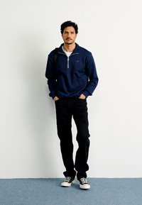Jūrinės tamsiai mėlynos zip-up megztinis su reljefine tekstūra, baltas marškinėliai, juodos kelnės ir juodai baltos sportbačiai; modelis stovi prieš paprastą foną.