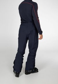 Personne portant un pantalon de ski bleu marine et des chaussures de ski noires, debout de dos face au spectateur sur un fond uni.