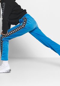 Blå träningbyxor med en svart sidstripe och ett upprepat logomönster, tillverkade av ett mjukt material med resårmidja.