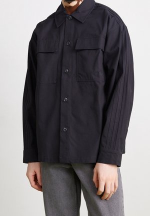 Homme portant une veste boutonnée noire avec deux poches poitrine et un pantalon gris, debout devant un fond uni.