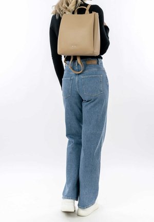 Femme portant un sac à dos marron clair, un haut à manches longues noir, un jean bleu et des baskets blanches, debout devant un fond blanc uni.