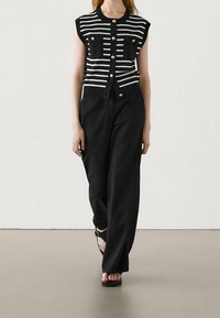 Haut sans manches à rayures noires et blanches, doté de deux poches frontales et de boutons, associé à un pantalon noir ample et des sandales noires.