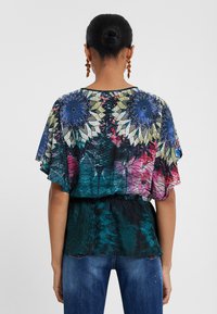 Blusa a fantasia floreale, multicolore, con maniche corte a volant. Presenta una vita cinchata e una combinazione di tonalità di blu, rosa e verde.