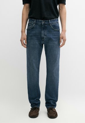 Mann trägt gerade geschnittene blaue Jeans, braune Schnürschuhe und ein schwarzes Kurzarmhemd vor einem schlichten weißen Hintergrund.
