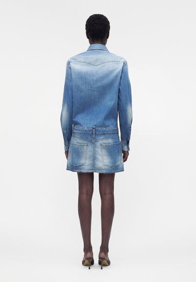 Άτομο που στέκεται με την πλάτη φοράει ένα ξεθωριασμένο μπλε τζιν μπουφάν και μια αντίστοιχη κοντή φούστα από τζιν με πίσω τσέπες και μπεζ παπούτσια με τακούνια.