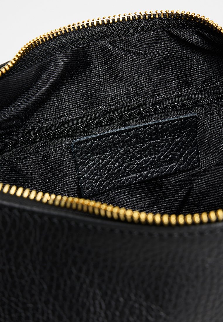 Intérieur d'un sac en cuir noir présentant une texture lisse, une fermeture éclair dorée et une petite étiquette en cuir, avec un revêtement en tissu noir.