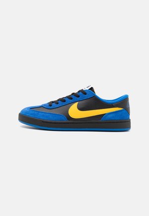Baskets basses en cuir noir, accents en daim bleu, logo Nike jaune, lacets noirs et semelle bleue sur fond blanc.