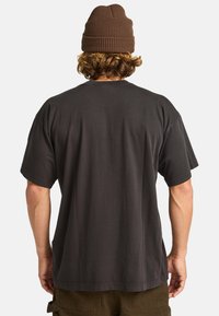 Mörkgrå kortärmad t-shirt med avslappnad passform, tillverkad av bomull, visad bakifrån, tillsammans med olivgröna cargo-byxor och en brun stickad mössa.