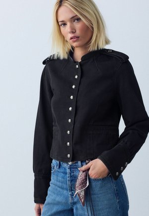 Giovane donna che indossa una giacca nera abbottonata e jeans blu con una sciarpa fantasia infilata nella tasca, su uno sfondo semplice.