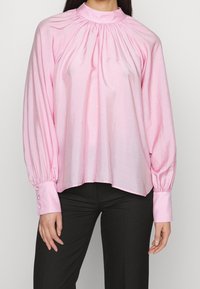 Blusa rosa a maniche lunghe con scollo arricciato, texture liscia e polsini aderenti con dettagli di bottoni. Abbinata a pantaloni neri.