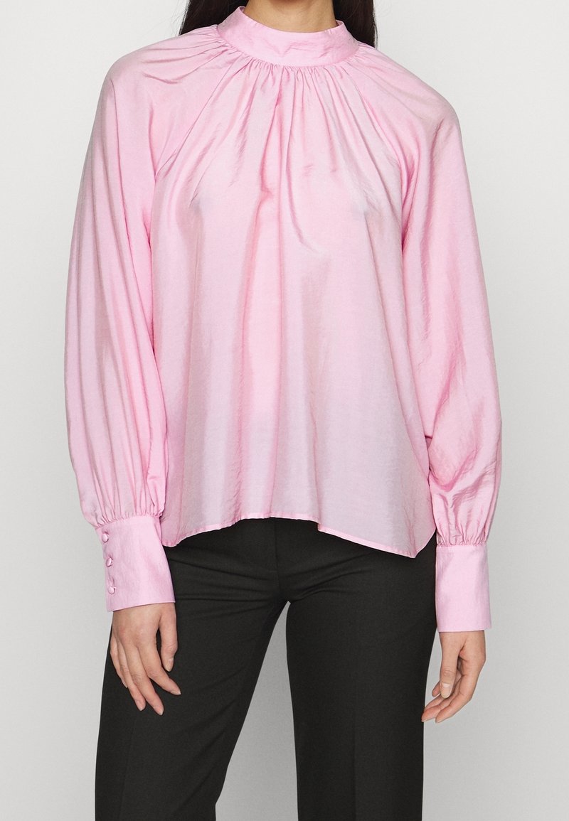 Blusa rosa a maniche lunghe con scollo arricciato, texture liscia e polsini aderenti con dettagli di bottoni. Abbinata a pantaloni neri.