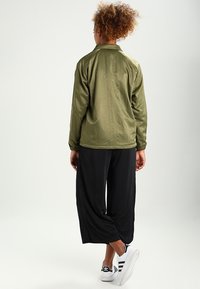 Mladý člověk s kudrnatými vlasy stojí čelem k odchodu, má na sobě olivově zelenou bundu, volné černé kalhoty a bílé tenisky Adidas.