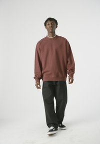 Bordeaux-roter Sweatshirt mit Rundhalsausschnitt und gerippten Bündchen, kombiniert mit lockeren schwarzen Hosen und schwarzen Segeltuchschuhen mit weißen Sohlen.