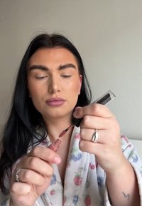 Applicateur de rouge à lèvres liquide retiré d'un tube transparent. Le produit est d'une teinte rose doux. L'arrière-plan est neutre avec un mur texturé.