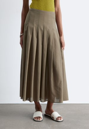 Femme portant une jupe midi plissée taille haute vert olive, un top jaune sans manches, des sandales blanches à enfiler, avec un bracelet et une bague en argent.