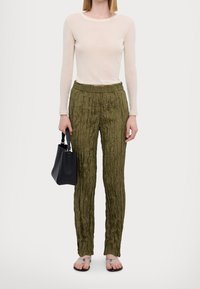 Vrouw in een doorzichtige beige blouse met lange mouwen, olijfgroene gekreukte broek, zilveren sandalen en een zwarte handtas aan haar zijde.