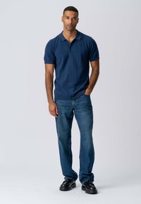 Mand iført en navy kortærmet skjorte med knapper, blå jeans og sorte sko, stående med den ene hånd i lommen foran en ensfarvet baggrund.