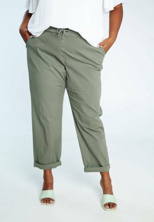 Personne portant un pantalon vert olive retroussé, un haut blanc et des sandales à talons ouvertes vert clair, debout les mains dans les poches.