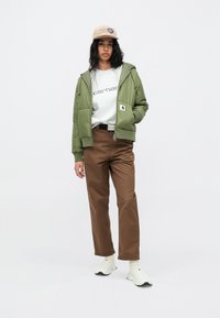 Olivno zelena jopica na zadrgo, sivi pulover z logotipom, rjave hlače, bele slip-on čevlje in bež kapa z logotipom in grafiko.