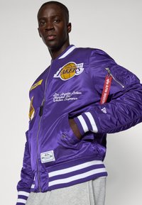 Fialový saténový bomber s brandingom Los Angeles Lakers, pruhovanými ribbed manžetami a viacerými vyšitými nášivkami. Detaily prispôsobiteľnej zipsovej vrecka.