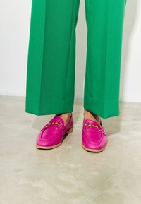 Roze leren instappers met decoratieve gouden hardware en een lichte zool, gecombineerd met groene wijde broekspijpen. Gladde textuur en levendige kleur.