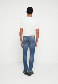 Vaqueros de mezclilla azul con corte de pierna recta, presentan un lavado claro, costuras visibles y una etiqueta de marca en la cintura trasera. Combinados con una camisa blanca.