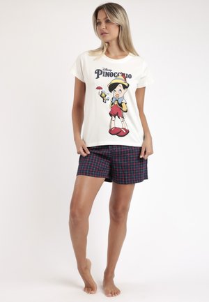 Mujer vistiendo camiseta blanca con gráfico de Pinocho y pantalones cortos de cuadros azul marino, de pie descalza contra un fondo claro y liso.