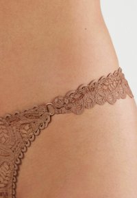 Spetsstring i en dämpad brun nyans med blommönster, med scalloped kanter och en liten metallbeslag.