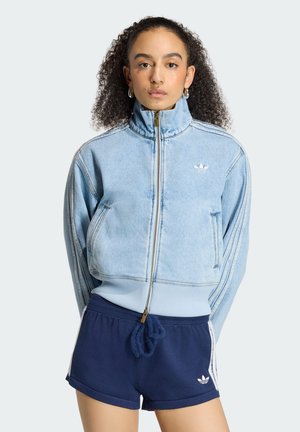 Donna con capelli ricci che indossa una giacca azzurro chiaro con zip e pantaloncini Adidas blu navy con strisce bianche e loghi.