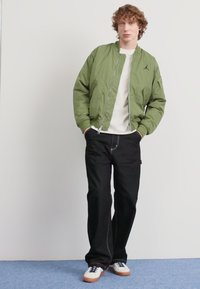Giacca bomber verde oliva, t-shirt bianca, jeans neri e sneaker basse con tomaia in pelle bianca e suola in gomma marrone; design semplice, vestibilità rilassata.