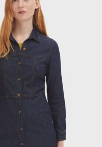 Vestido camisero de mezclilla oscura con botones en la parte delantera, mangas largas, bolsillos en el pecho y detalles de costura en contraste naranja. Textura suave, forma ajustada.