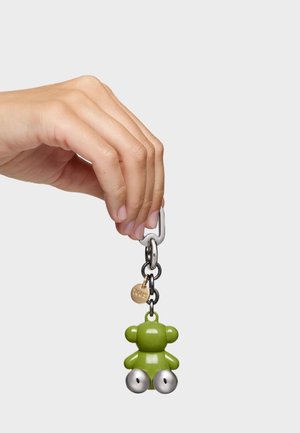 Mano sosteniendo un llavero con un charm en forma de oso verde y dos campanas de plata adjuntas.