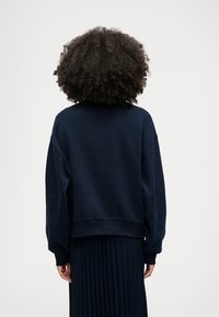 Ναυτικό μπλε φούτερ με χαλαρή εφαρμογή, ριμπ μανσέτες και στρίφωμα. Διαθέτει στρογγυλή λαιμόκοψη και χαμηλούς ώμους. Μαλακή υφή υφάσματος.